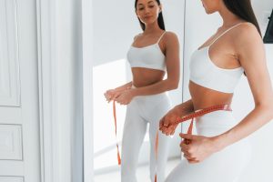 Uses measuring tape. Young caucasian woman with slim body shape is indoors at daytime Uses measuring tape. Young caucasian woman with slim body shape is indoors at daytime.